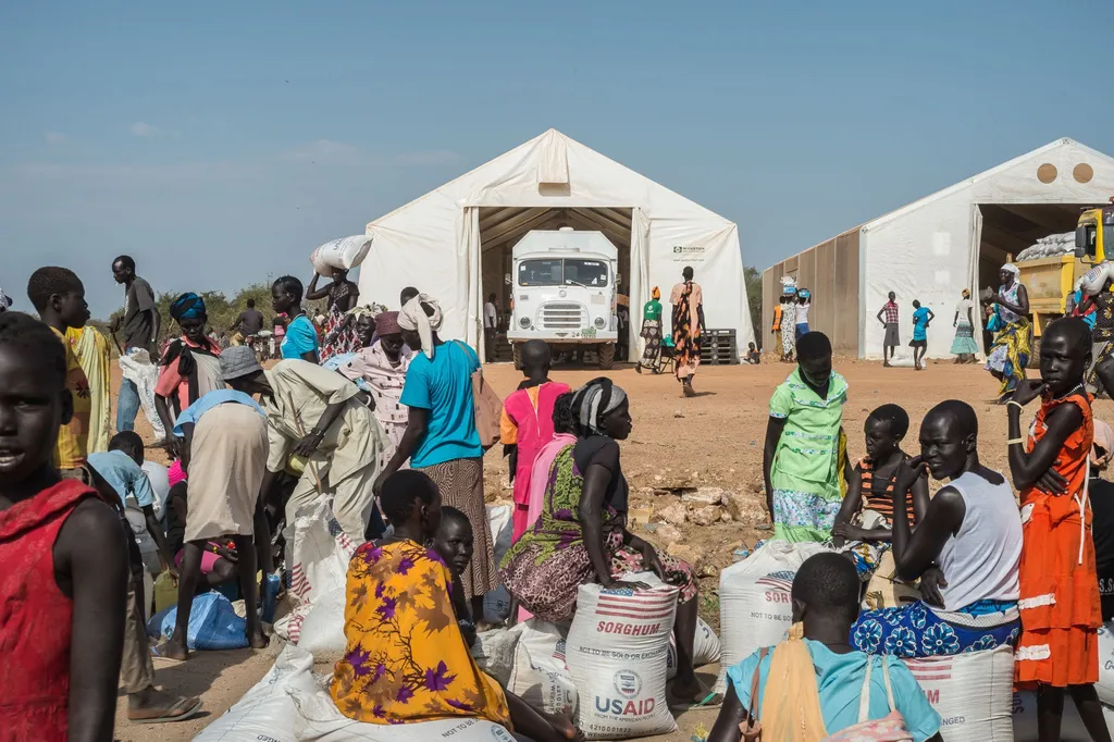 A crowded relief camp in Sudan with people waiting near large tents and USAID bags of sorghum, with a truck parked in the background.