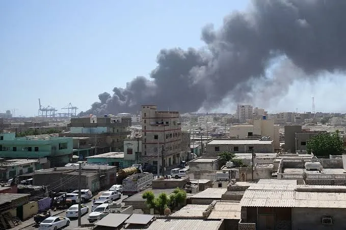 A wide aerial view of a crowded city with dense black smoke billowing into the sky from an industrial area or building fire in the distance.