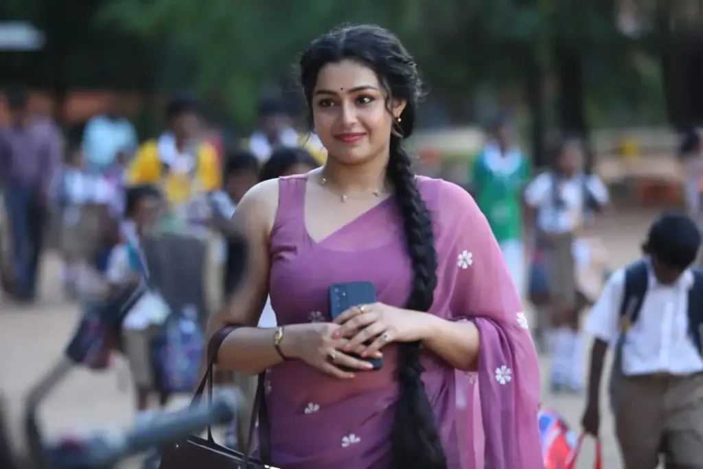 Actress Mokksha wearing a mauve saree with white floral patterns, holding a smartphone and handbag, with school children walking in the blurred background.