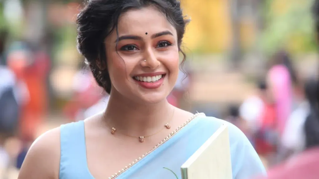 Close-up portrait of actress Mokksha smiling warmly while wearing a light blue sleeveless blouse and matching saree, with her hair styled in an elegant updo and holding a book in an outdoor setting.