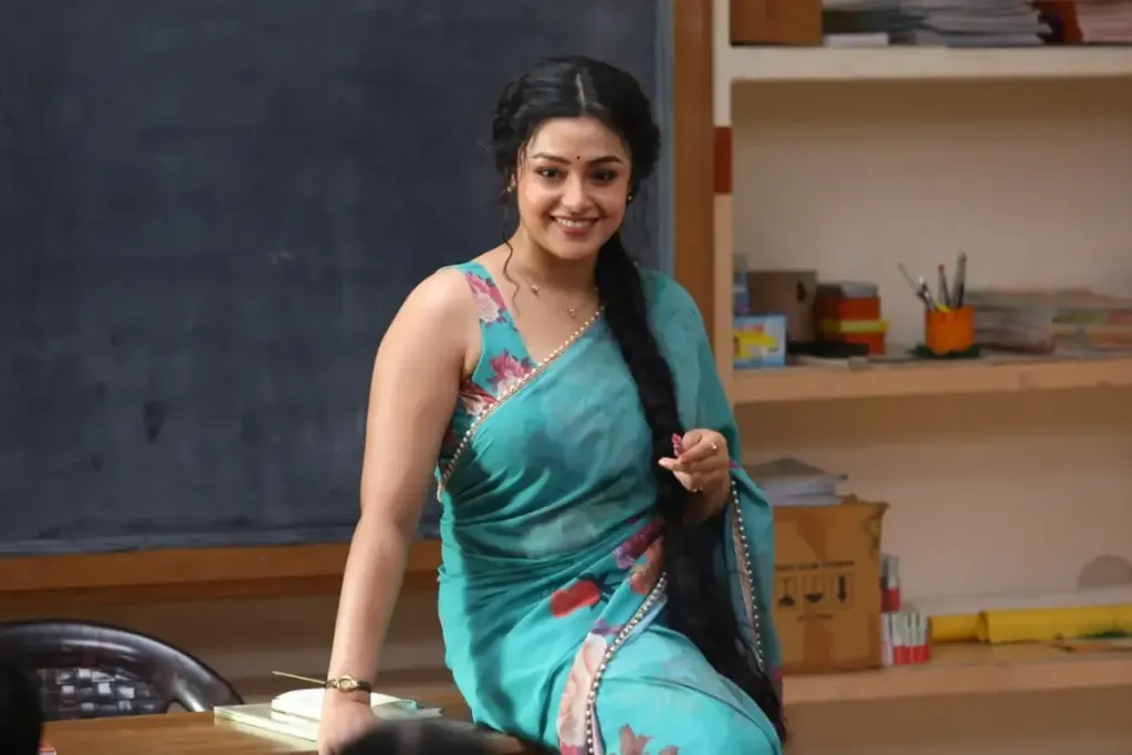 Actress Mokksha portraying Meenu Miss in a classroom setting, wearing a turquoise floral saree with her long dark hair in a single braid, smiling while leaning against a desk.