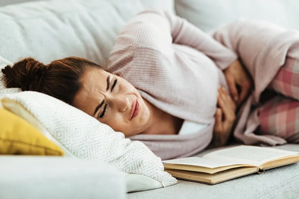 A woman lying on a light grey couch with her eyes squeezed shut in pain, holding her stomach with her hand while an open book rests nearby.