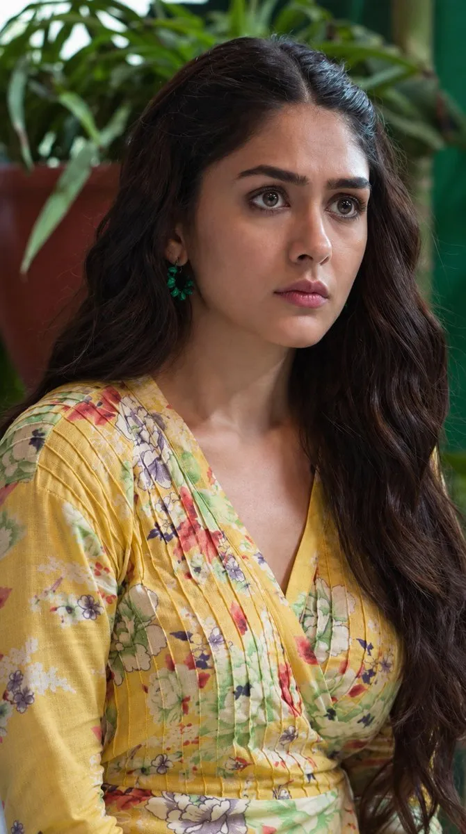 Close-up of actress Mrunal Thakur wearing a yellow floral wrap top with green earrings, looking intently to the side with a serious expression against a blurred green plant background.